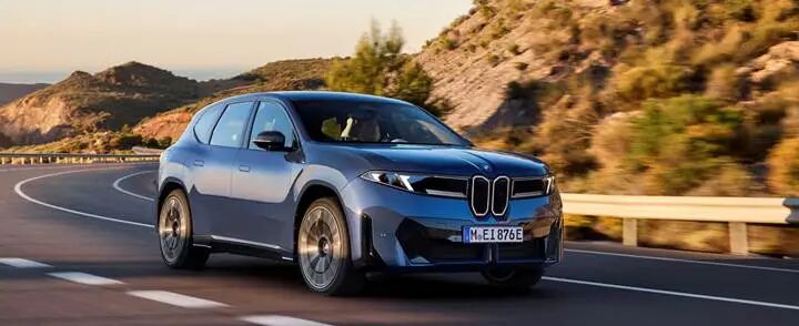 A blue BMW iX3 drives along a winding road surrounded by hills and trees.